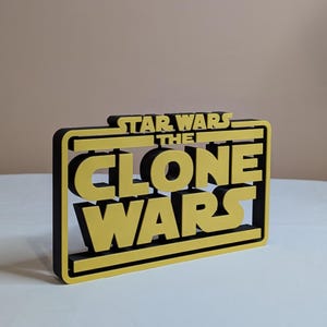 Könnte beinhalten: Ein schwarz-gelbes 3D-gedrucktes Schild mit der Aufschrift "Star Wars The Clone Wars".