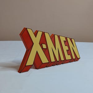 May include: A 3D printed sign that reads "X-MEN" in yellow letters with black outlines on a red background.