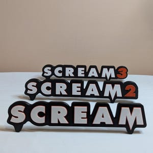 Könnte beinhalten: Drei schwarze und weiße Kunststoffschilder mit dem Wort "SCREAM" in weißen Buchstaben mit orangefarbenem Umriss. Die Schilder sind übereinander gestapelt, wobei das oberste Schild "SCREAM 3", das mittlere Schild "SCREAM 2" und das unterste Schild "SCREAM" lautet.