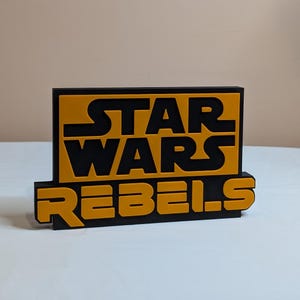 Könnte beinhalten: Ein schwarzes und gelbes Schild mit dem Text "STAR WARS REBELS" in fetten Blockbuchstaben.