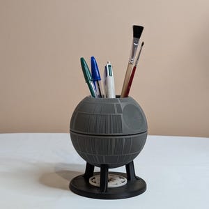 Può includere: Un portaoggetti da scrivania grigio a forma di Morte Nera con una base nera e quattro gambe. L'organizzatore è pieno di penne e pennelli.