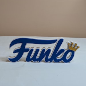 Peut inclure: Un présentoir Funko Pop blanc avec le mot "Funko" en lettres bleues et une couronne dorée sur le côté droit.