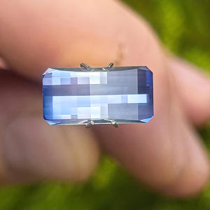 Peut inclure: Une pierre précieuse bleue rectangulaire à facettes avec un aspect clair et transparent. La pierre présente une bande horizontale distincte de couleur bleu plus clair au centre, maintenue par de petites griffes métalliques fines.