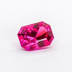 May include: A vibrant, hot pink gemstone with a rectangular, faceted cut. The stone features sharp, geometric edges and a highly reflective surface that creates deep, saturated colour variations and bright light refractions throughout its structure.