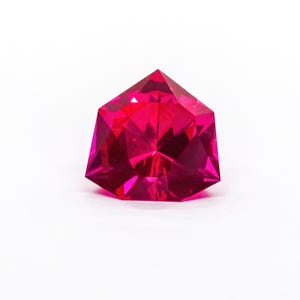Puede incluir: Una gema de color rosa intenso y vibrante con un corte geométrico facetado. La piedra presenta una parte superior puntiaguda y una forma angular simétrica con numerosas superficies reflectantes que crean un aspecto brillante y centelleante.