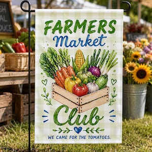 Op de afbeelding: Een rechthoekige tuinvlag met een lichtgroene ruitjesachtergrond. Het toont een houten kist gevuld met verse groenten, waaronder wortels, maïs, paprika's en tomaten. De tekst luidt FARMERS Market Club en WE CAME FOR THE TOMATOES.