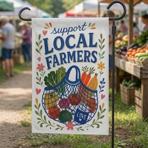 Op de afbeelding: Een witte tuin vlag met de tekst support LOCAL FARMERS in het blauw, voorzien van een kleurrijke illustratie van een nettas gevuld met verse producten zoals wortels, tomaten en bladgroenten, versierd met kleine hartjes en bloemmotieven.