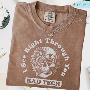 Może przedstawiać: Brązowa koszulka w kolorze espresso z grafiką przedstawiającą czaszkę, kwiaty i napis "I See Right Through You RAD TECH". Srebrny naszyjnik z wisiorkiem spoczywa na koszulce. Koszulka wykonana jest z materiału Comfort Colors.