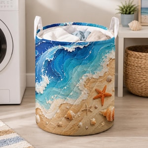 Könnte beinhalten: Ein zylindrischer Wäschekorb mit einem Druckmotiv von Meereswellen, die auf einen Sandstrand treffen. Die Außenseite zeigt blaues und türkisfarbenes Wasser mit weißem Schaum, während der Sand mit einem leuchtend orangefarbenen Seestern und Muscheln verziert ist.