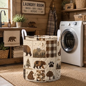 Könnte beinhalten: Ein zylindrischer Wäschekorb mit einer gesteppten Stoffaußenseite im Patchwork-Design. Das Muster zeigt braune Bären, Tannenbäume, Berglandschaften und karierte Quadrate in Beige-, Braun- und Cremetönen mit weißen Griffen.