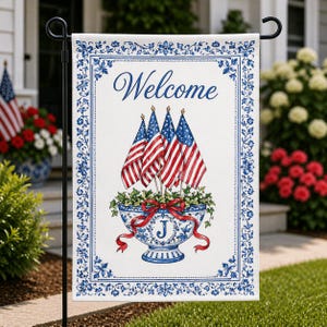 May include: A white garden flag with a blue floral border and the word "Welcome" in script. The flag features a blue and white bowl with American flags and a red bow. The initial "J" is on the bowl.