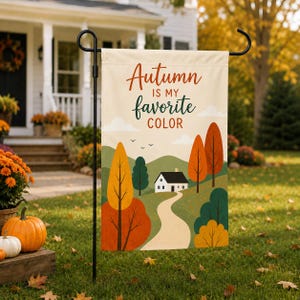 Puede incluir: Una bandera de jardín rectangular colgada en un soporte de metal negro. La bandera presenta un fondo crema con el texto "Autumn IS MY favorite COLOR" en marrón y verde. Representa una casa blanca, un camino sinuoso y árboles.