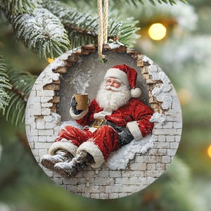Op de afbeelding: Ronde kerstversiering met de kerstman in een rood pak en muts, met een mok in zijn hand. De versiering heeft een bakstenen muurontwerp en hangt aan een touwtje. De achtergrond is een vage kerstboom.