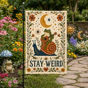 May include: A rectangular garden flag with a cream background featuring a central illustration of a green frog playing a guitar while sitting on a blue snail. The design includes a crescent moon, stars, an eye, and the text STAY WEIRD in blue.