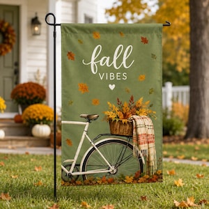 Peut inclure: Un drapeau de jardin rectangulaire sur fond vert sauge avec le texte blanc fall VIBES et un petit cœur. Le motif représente un vélo blanc avec un panier en osier rempli de feuilles d'automne, une couverture à carreaux et des feuilles tombantes.