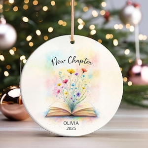 May include: A circular Christmas ornament with the text "New Chapter" and "Olivia 2025". The design includes an open book with colourful flowers and a watercolour background. It is suspended by a gold string.