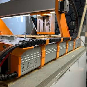 May include: A CNC machine with a silver and orange frame. The machine has a long, rectangular base with a series of orange brackets. A black cable chain runs along the side. The machine is set on a wooden surface.