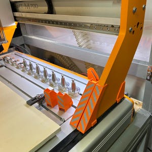 Può includere: Una macchina CNC arancione e argento con un pannello di legno chiaro in posizione. La macchina ha diverse punte argentate e supporti arancioni con strisce a zig-zag riflettenti. Il telaio della macchina è argento e arancione.