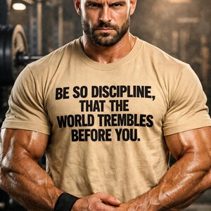Puede incluir: Una camiseta color beige con el texto "BE SO DISCIPLINE, THAT THE WORLD TREMBLES BEFORE YOU." La persona de la imagen tiene el pelo corto y barba. La persona está en un gimnasio.