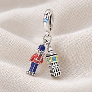 Puede incluir: Un charm de plata con un guardia británico colgante con uniforme rojo y azul junto a una torre del reloj Big Ben en miniatura con detalles en amarillo y azul. El pasador superior está engastado con una pequeña piedra de cristal azul claro.