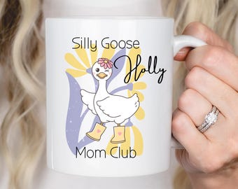Peut inclure: Tasse en céramique blanche avec un dessin de canard en dessin animé portant des bottes jaunes et une fleur. Le texte "Silly Goose Holly Mom Club" est écrit en noir, avec des accents floraux jaunes et violets.