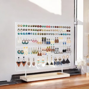 May include: A white metal earring display stand with a rectangular wooden base, featuring multiple horizontal bars for organizing various styles of earrings, including studs, hoops, and dangling designs in diverse colors, shapes, and materials.