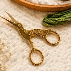May include: Gold-colored metal embroidery scissors shaped like a crane with a polished finish. The handles form the bird's body and wings, while the sharp, pointed blades extend from the beak. The scissors feature intricate engraved details.