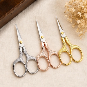 May include: Three pairs of small embroidery scissors with ornate, embossed handles in silver, rose gold, and gold finishes. Each pair features sharp, pointed steel blades and a central screw, showcasing intricate floral patterns on the metal handles.