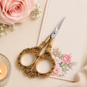 May include: Gold-colored metal embroidery scissors featuring ornate, intricate filigree handles and sharp, polished silver-toned steel blades. The scissors are designed with oval finger loops and a central pivot screw, showcasing a detailed decorative pattern.