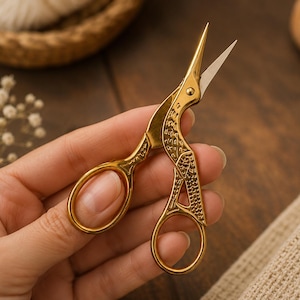 May include: Gold-colored metal embroidery scissors shaped like a crane. The scissors feature a polished finish with intricate engraved feather detailing along the handles and blades, ending in sharp, pointed tips for precise cutting tasks.