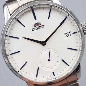 May include: Close-up of an Orient watch with a white face, black hands, and a date display at the 3 o'clock position. The watch has a silver stainless steel band and a small seconds subdial. The Orient logo is visible.