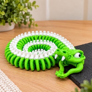 May include: A coiled articulated snake toy featuring a bright green body with a white spine-like ridge along the top. The snake has a detailed head with white fangs, a wide mouth, and a single visible large white eye with a black pupil.