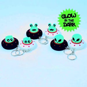May include: A collection of alien-themed keychains. Each keychain features a green alien figure atop a black and white UFO with a silver keyring. The keychains are arranged on a light blue surface, with a green starburst sign that reads "GLOW IN THE DARK".