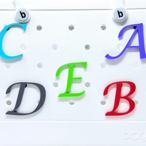 May include: A white tote bag with colorful letter charms spelling out "C A D E B". The letters are in blue, purple, green, gray, and red. The bag has a perforated design and the brand name "bogg" is visible.