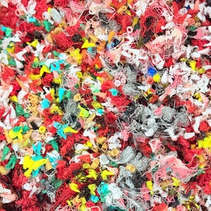 May include: A dense collection of tangled 3D printing filament waste in a vibrant array of colors including red, white, yellow, black, green, and pink. The material consists of thin, coiled, and knotted plastic strands creating a textured surface.