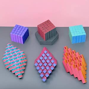 May include: An assortment of colorful geometric puzzles in various shapes. The puzzles include cubes in purple, red, and blue, and diamond-shaped structures in pink, blue, and orange. The background is a combination of pink and gray.