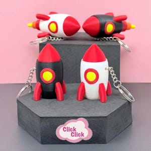 May include: Four rocket-shaped keyrings in red, black, and white. Each keyring has a red cone top, a yellow circle, and red fins. The keyrings are on a grey platform with a pink background. The text "Click Click" is on a sticker.