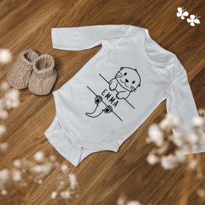 Könnte beinhalten: Weißer langärmliger Baby-Strampler mit einer schwarz-weißen Illustration eines Otters, der an einem Ast hängt. Der Strampler trägt den Namen "EMMA" aufgedruckt.