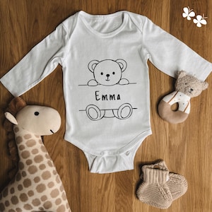 Puede incluir: Mono blanco de manga larga para bebé con una ilustración en blanco y negro de un osito de peluche asomándose por encima de una pared con el nombre "Emma" impreso debajo.