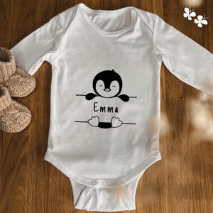 Könnte beinhalten: Weißer langärmliger Baby-Strampler mit einem schwarzen Pinguin-Motiv, der ein Schild mit der Aufschrift "Emma" hält.