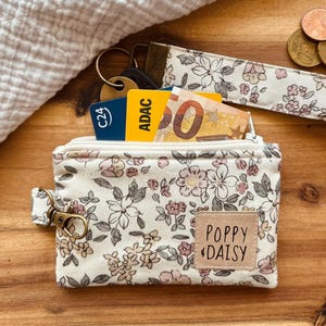 Könnte beinhalten: Eine kleine, floral gemusterte Geldbörse mit Reißverschluss, die Karten und Bargeld enthält. Die Geldbörse ist cremefarben mit rosa und grauen Blumenmustern. Ein passender Handgelenkriemen und Schlüssel sind sichtbar. Das Etikett auf der Geldbörse lautet "POPPY DAISY."