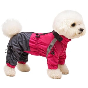 Puede incluir: Un perro con un mono impermeable rosa y gris. El mono tiene una tira reflectante en las mangas y las piernas, y un cuello negro. El perro es un Bichon Frise blanco, de pie sobre un fondo blanco.