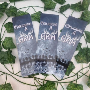 May include: Three dark blue bookmarks with the text "Conjuring A Grim" in white. The bookmarks feature a misty forest scene with flying birds and deer antlers. Green ivy and leaves are in the background.