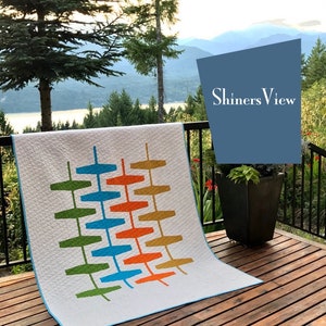 May include: A white quilt with a geometric pattern in shades of green, blue, orange, and yellow. The quilt is folded and placed on a wooden deck with a view of mountains and a lake in the background. The text "Shiners View" is visible in the upper right corner of the image.