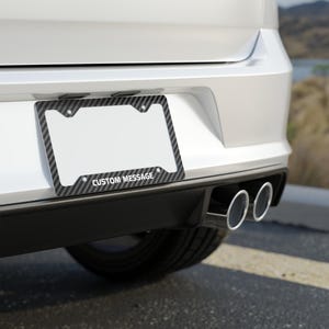 May include: A carbon fiber license plate frame with the text "CUSTOM MESSAGE" in white lettering. The frame is mounted on the back of a white car, with dual exhaust pipes visible below.
