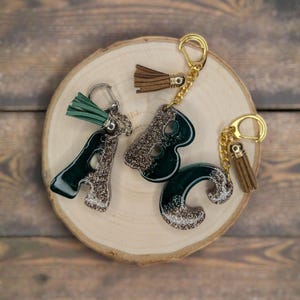 May include: Three letter keychains spelling "ABC" are displayed on a wooden slice. The letters are dark green with silver glitter accents. Each keychain has a gold-colored clasp and a tassel. The "A" has a green tassel, while the "B" and "C" have brown tassels.