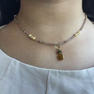 May include: A gold beaded choker necklace with a small pineapple charm. The necklace is made with light brown and gold beads.