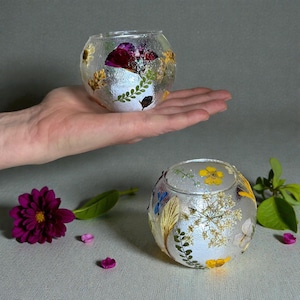 May include: Two clear, round glass candle holders decorated with dried flowers and leaves. One is held in a hand, the other sits on a surface. A purple flower and petals are in the foreground. The candle holders are a unique home decor item.