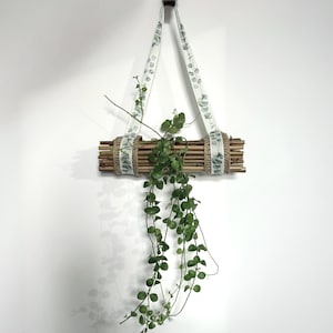 Könnte beinhalten: Ein hängender Pflanztopf aus Bambus und geflochtenem Bindfaden, aufgehängt an einem weißen Stoffband mit grünem Blättermuster. Der Pflanztopf enthält eine grüne Hängepflanze, die ein natürliches Dekorationselement schafft.