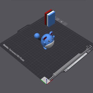 May include: A 3D-printed model of a blue and white cartoon character with a round body, large ears, and a tail. A small, rectangular block with red, white, and blue stripes is also present. The objects are on a dark grey surface.